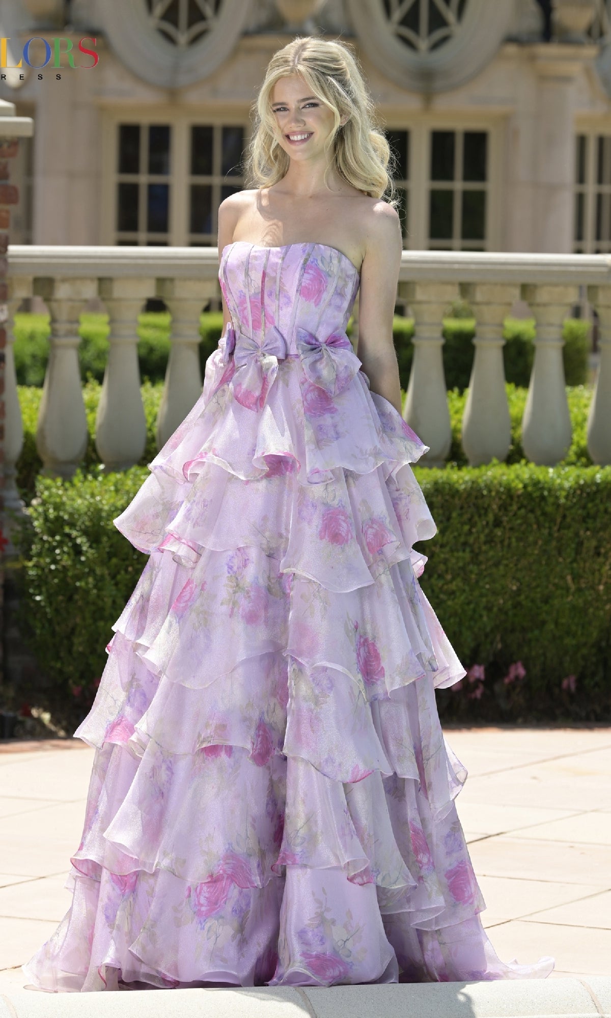 A woman stands outdoors wearing the Colors Dress 3809, a strapless long prom dress with a corset bodice and floor-length pastel purple floral gown featuring multiple ruffled layers.